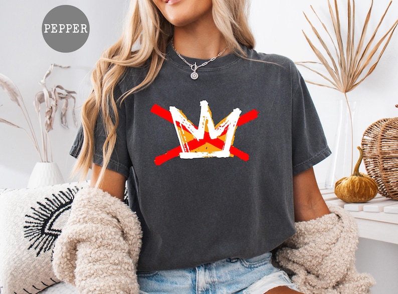 May include: Dark gray t-shirt featuring a graphic of a crown with red lines through it. The crown is white, yellow, and orange. The word "PEPPER" is in a gray circle in the upper left corner. The shirt is a casual style.