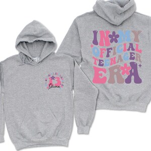 May include: Gray hooded sweatshirt with the text "In My Official Teenager Era" in colorful retro lettering on the back and a flower design on the front.