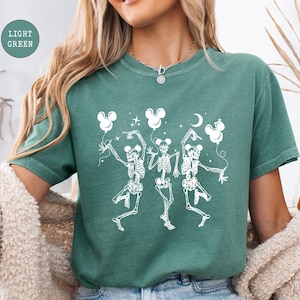 May include: A light green t-shirt featuring a whimsical design of three dancing skeletons holding Mickey Mouse-shaped balloons. The skeletons are white, and the balloons, stars, and moon are also white. The shirt has a relaxed fit.