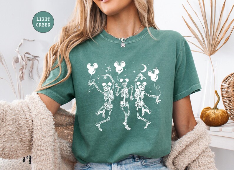 May include: A light green t-shirt featuring a whimsical design of three dancing skeletons holding Mickey Mouse-shaped balloons. The skeletons are white, and the balloons, stars, and moon are also white. The shirt has a relaxed fit.