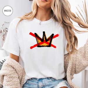 May include: White t-shirt featuring a graphic of a crown with red cross-out lines. The crown design has black, yellow, and orange accents. The word "WHITE" is in a white circle in the upper left corner. The shirt is a casual, everyday wear item.