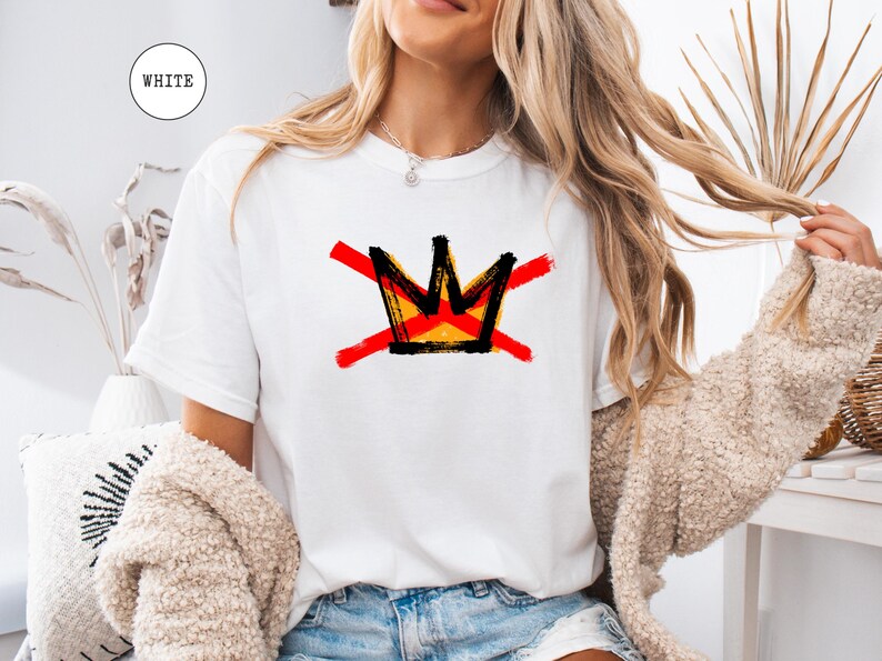 May include: White t-shirt featuring a graphic of a crown with red cross-out lines. The crown design has black, yellow, and orange accents. The word "WHITE" is in a white circle in the upper left corner. The shirt is a casual, everyday wear item.