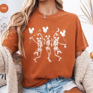May include: Burnt orange t-shirt featuring a white graphic of three dancing skeletons holding Mickey Mouse-shaped balloons. The skeletons are in a playful pose, with a crescent moon and stars. The shirt has a crew neck.