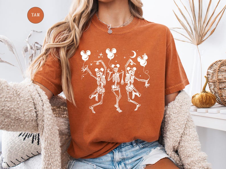 May include: Burnt orange t-shirt featuring a white graphic of three dancing skeletons holding Mickey Mouse-shaped balloons. The skeletons are in a playful pose, with a crescent moon and stars. The shirt has a crew neck.