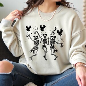 May include: Cream-colored sweatshirt featuring a black graphic of three dancing skeletons holding Mickey Mouse-shaped balloons. The design includes a crescent moon and stars. The sweatshirt has ribbed cuffs and a crew neck.