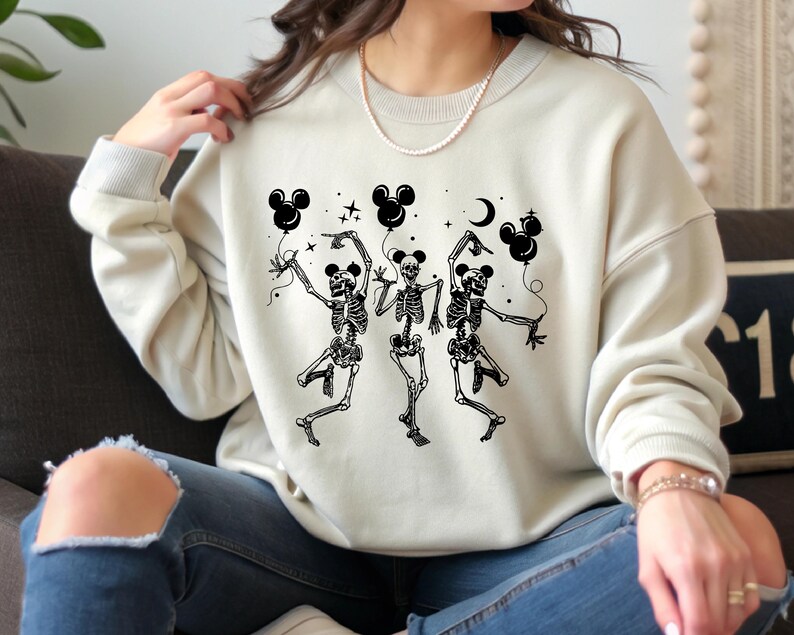 May include: Cream-colored sweatshirt featuring a black graphic of three dancing skeletons holding Mickey Mouse-shaped balloons. The design includes a crescent moon and stars. The sweatshirt has ribbed cuffs and a crew neck.