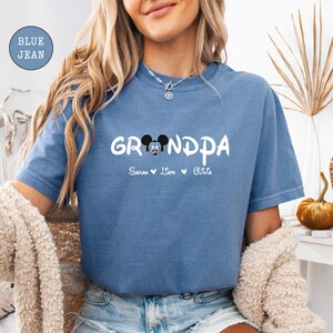 Op de afbeelding: Een jeansblauw T-shirt met de tekst "Grandpa" in wit met een Mickey Mouse-kopgrafiek en de namen "Sarah, Liam, Olivia" eronder.