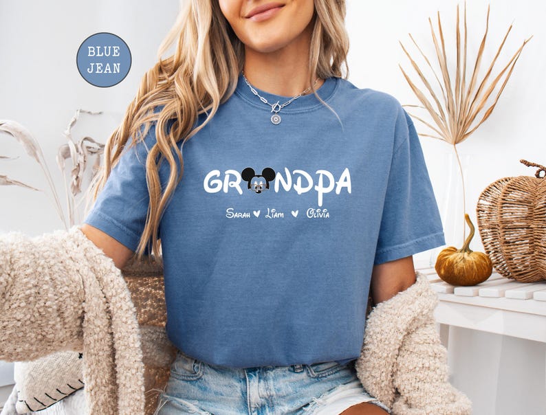 Op de afbeelding: Een jeansblauw T-shirt met de tekst "Grandpa" in wit met een Mickey Mouse-kopgrafiek en de namen "Sarah, Liam, Olivia" eronder.