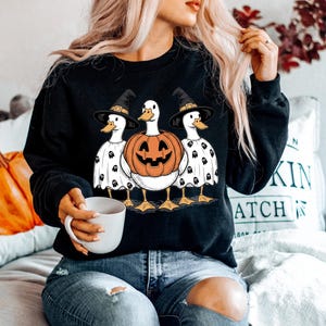 Peut inclure: Sweat-shirt noir avec un motif d'Halloween repr&eacute;sentant trois canards. Le canard central est une citrouille sculpt&eacute;e, flanqu&eacute;e de canards portant des chapeaux de sorci&egrave;re et des costumes de fant&ocirc;mes. Les canards ont des pieds et des becs jaunes.