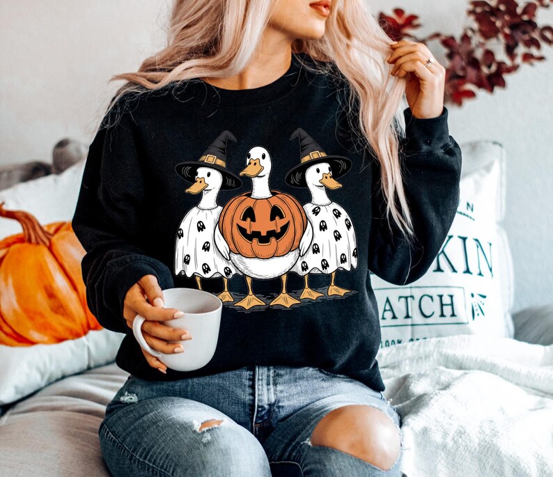 Peut inclure: Sweat-shirt noir avec un motif d'Halloween repr&eacute;sentant trois canards. Le canard central est une citrouille sculpt&eacute;e, flanqu&eacute;e de canards portant des chapeaux de sorci&egrave;re et des costumes de fant&ocirc;mes. Les canards ont des pieds et des becs jaunes.