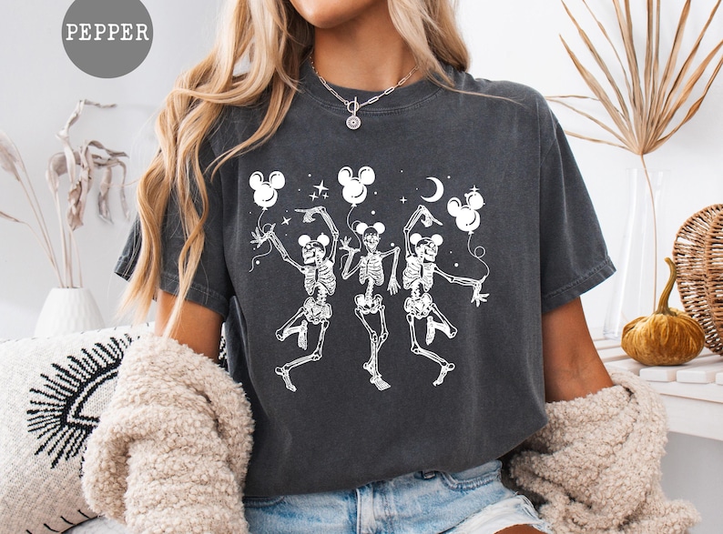May include: Dark gray t-shirt featuring a white graphic of three dancing skeletons holding Mickey Mouse-shaped balloons. The design includes a crescent moon and stars. The word "PEPPER" is in a circle at the top left.