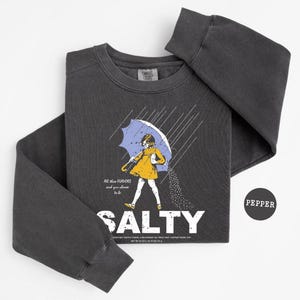 May include: Dark gray sweatshirt with a graphic of a person under a blue umbrella, with the word "SALTY" in white. The text "All these flavors and you choose to be" is also on the shirt. A black circle with the word "PEPPER" is on the right.