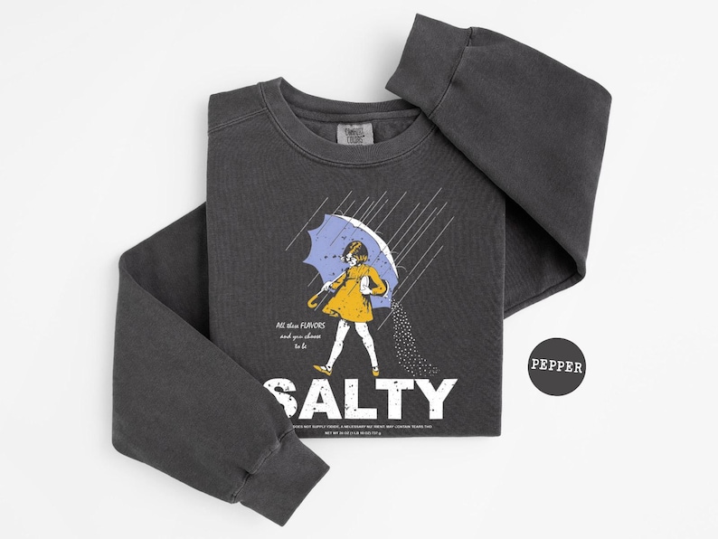May include: Dark gray sweatshirt with a graphic of a person under a blue umbrella, with the word "SALTY" in white. The text "All these flavors and you choose to be" is also on the shirt. A black circle with the word "PEPPER" is on the right.