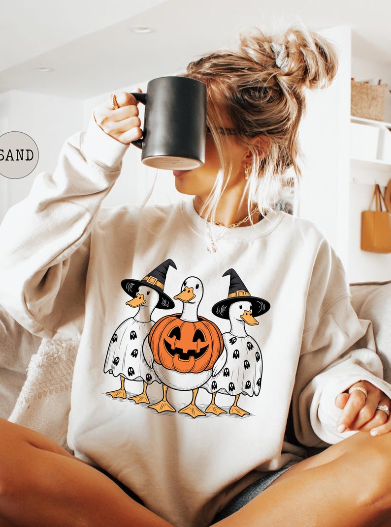 Peut inclure: Sweat-shirt cr&egrave;me avec un motif d'Halloween. Le motif pr&eacute;sente trois canards, deux portant des chapeaux de sorci&egrave;re et des capes fant&ocirc;mes, et un avec un corps de citrouille sculpt&eacute;e. Les canards ont des becs et des pattes oranges.