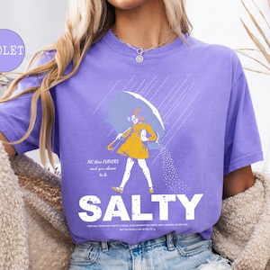 May include: Violet-colored t-shirt with a graphic of a person walking under an umbrella, with the word "SALTY" in large white letters. The graphic includes text that reads "All these flavors and you choose to be".