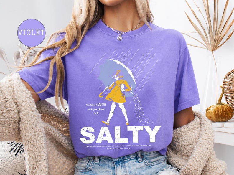 May include: Violet-colored t-shirt with a graphic of a person walking under an umbrella, with the word "SALTY" in large white letters. The graphic includes text that reads "All these flavors and you choose to be".