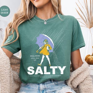 May include: Light green t-shirt with a graphic of a person under a purple umbrella in the rain. The person is wearing a yellow dress and is sprinkling salt. The word "SALTY" is printed in large white letters below the graphic.