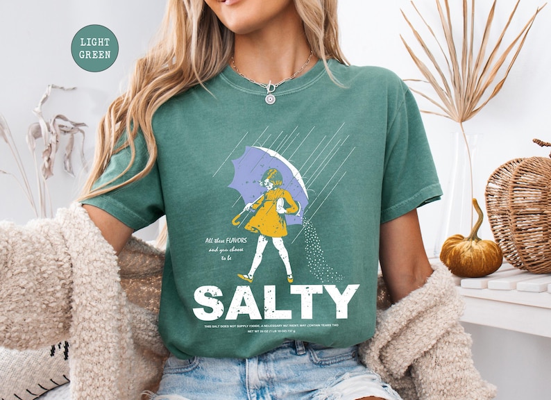 May include: Light green t-shirt with a graphic of a person under a purple umbrella in the rain. The person is wearing a yellow dress and is sprinkling salt. The word "SALTY" is printed in large white letters below the graphic.