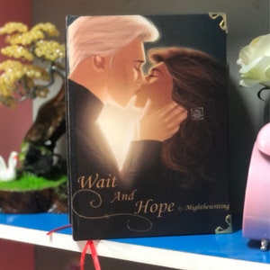 May include: A black hardback book with a gold-coloured design. The cover features a couple kissing with a glowing light behind them. The title of the book is "Wait And Hope" by Mightbewriting.