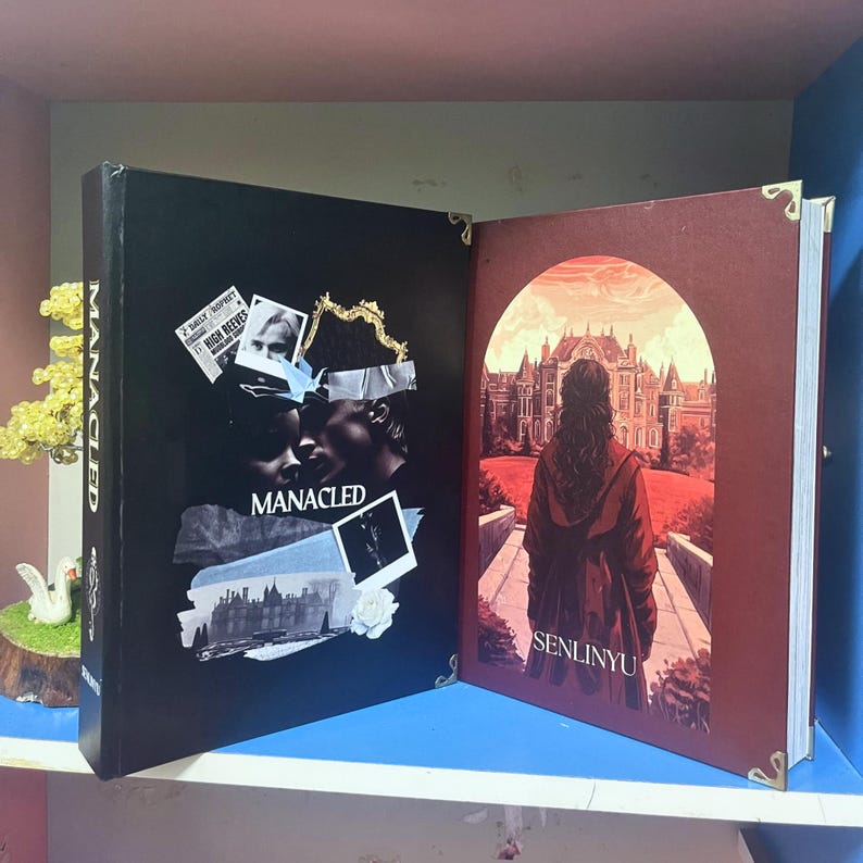 May include: An open book with a black cover featuring the word "MANACLED" and a collage of images. The right page shows a person in a red coat facing a building, with the word "SENLINYU" below. Gold corner protectors are visible.
