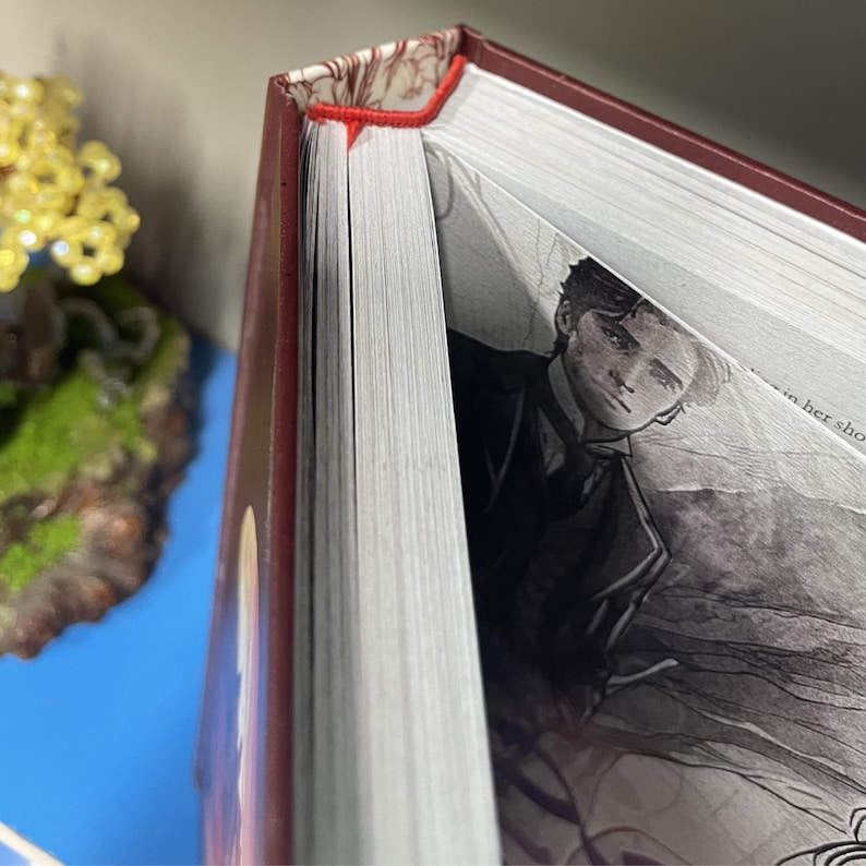 May include: A hardcover book with a burgundy cover and a red bookmark. The pages are white, and the book is open to a black and white illustration of a person in a suit. The book's spine is visible, showing the pages' edges.