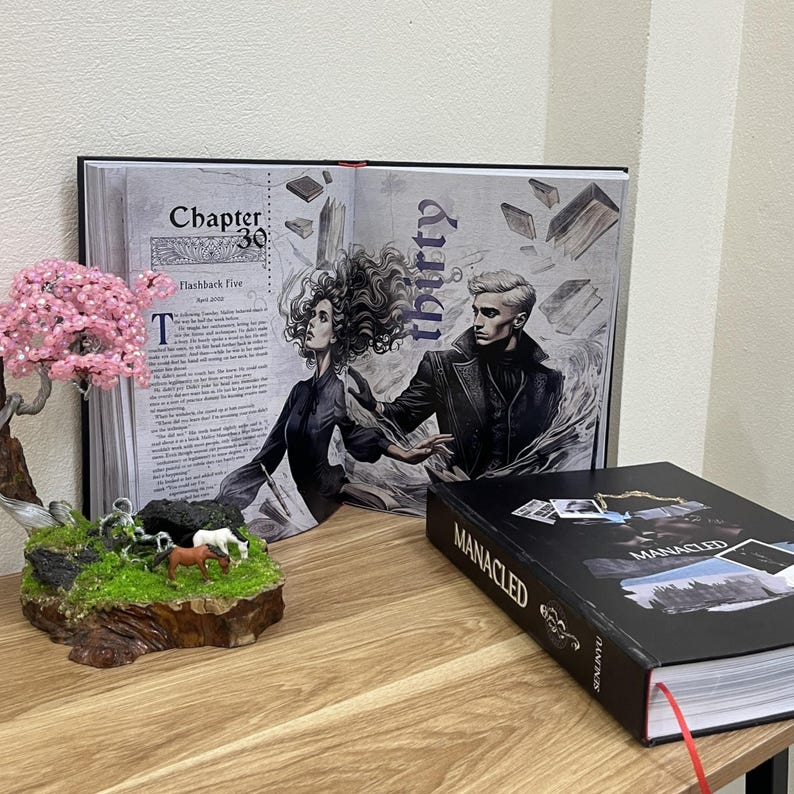 May include: An open book with a detailed illustration of two figures, with the word "thirty" and "Chapter 30" visible. A closed book with the title "MANACLED" is on the right. A small decorative bonsai tree and a miniature landscape are in the foreground.