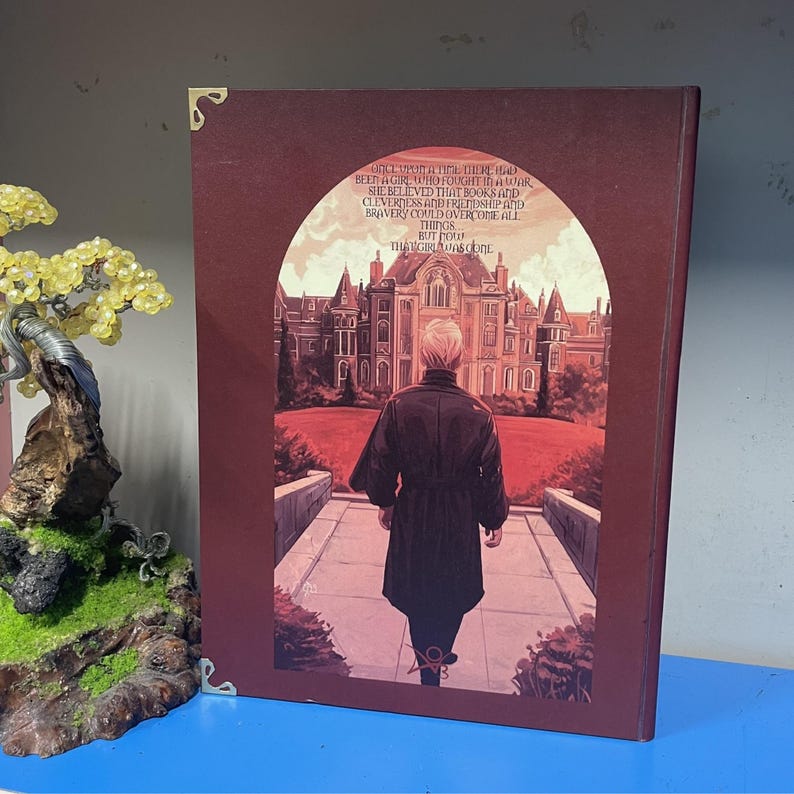 May include: A burgundy-colored book cover with a printed illustration. The image depicts a person walking towards a large building. Text on the cover reads, "ONCE UPON A TIME THERE HAD BEEN A GIRL WHO FOUGHT IN A WAR..."