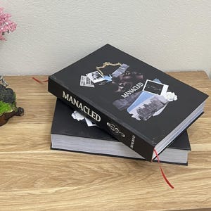 May include: Two black books stacked on a wooden surface. The top book has the word "MANACLED" in white, with a collage of images. A red ribbon bookmark extends from the top book.