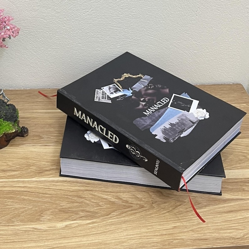 May include: Two black books stacked on a wooden surface. The top book has the word "MANACLED" in white, with a collage of images. A red ribbon bookmark extends from the top book.