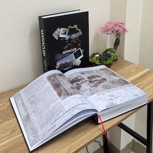 May include: Two hardcover books with the title "MANACLED" are displayed. One is open, revealing detailed illustrations and text. The cover features a collage of images and the title. A red ribbon marks a page. A decorative bonsai tree sits nearby.