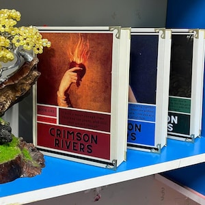 May include: Three books with white spines and metal corner protectors. The first book, "Crimson Rivers," has a red cover with a hand holding a flaming heart. The other two books have blue and black covers.