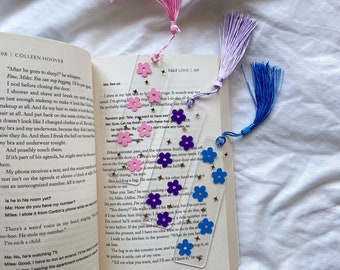 Peut inclure: Un marque-page en acrylique transparent avec un motif de fleurs roses, violettes et bleues. Le marque-page a un pompon en haut et des accents de paillettes dorées. Le marque-page est sur un livre avec le texte "Ugly Love 109" visible.