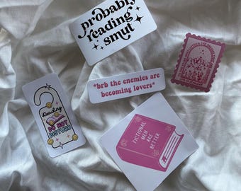 Peut inclure: Plusieurs autocollants blancs avec du texte noir et rose. L'un d'eux dit "probably reading smut". Un autre dit "*brb the enemies are becoming lovers*". Un autocollant rose représente un livre et dit "Fictional Men Are Better."