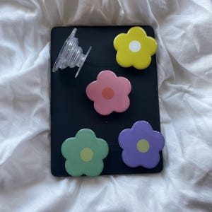 Peut inclure: Quatre poign&eacute;es de t&eacute;l&eacute;phone en forme de fleur color&eacute;es sur un fond noir. Les poign&eacute;es sont jaunes, roses, vertes et violettes. Une poign&eacute;e de t&eacute;l&eacute;phone transparente est &eacute;galement visible.