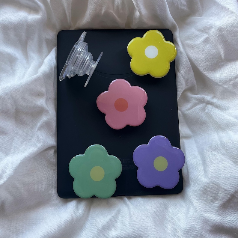 Peut inclure: Quatre poign&eacute;es de t&eacute;l&eacute;phone en forme de fleur color&eacute;es sur un fond noir. Les poign&eacute;es sont jaunes, roses, vertes et violettes. Une poign&eacute;e de t&eacute;l&eacute;phone transparente est &eacute;galement visible.