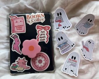 Peut inclure: Une coque de téléphone transparente avec divers autocollants. Les autocollants comprennent une fleur rose, une axolotl rose, un dinosaure rose, un livre rose avec le texte "Emotional Support Kindle", un ruban rose avec le texte "Hot Girl Book Club", et un autocollant rose avec le texte "Books & Coffee". Il y a aussi plusieurs autocollants de fantômes blancs avec des livres dans leurs mains.