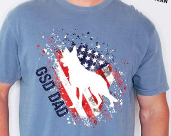May include: Blue short-sleeve t-shirt with a graphic featuring a white silhouette of a German Shepherd Dog (GSD) over a stylized American flag design. The text "GSD DAD" is printed on the shirt. The text "SHOWN IN BLUE JEAN" is also printed on the shirt.
