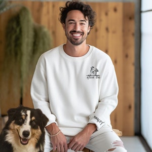 May include: A white sweatshirt with a black and white illustration of an Australian Shepherd dog peeking over the top. The text "Aussie Dad" is printed below the dog. The sweatshirt has a black cuff with the name "Harley" printed on it.