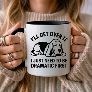 May include: White ceramic mug with a black interior and handle. The mug features a black and white illustration of a basset hound and the text "I'll Get Over It, I Just Need To Be Dramatic First."
