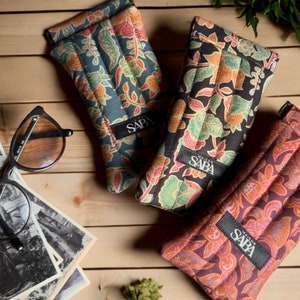 May include: Three quilted eyeglass cases with floral patterns. The cases are in shades of blue, black, and brown. Each case has a black label with the text "Maison de Saba" printed on it.