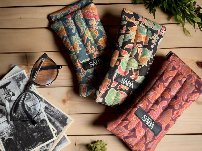 May include: Three quilted eyeglass cases with floral patterns. The cases are in shades of blue, black, and brown. Each case has a black label with the text "Maison de Saba" printed on it.