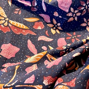 May include: A dark blue fabric with a repeating pattern of pink, orange, and white floral designs. The fabric is folded and draped, showing the intricate details of the pattern.