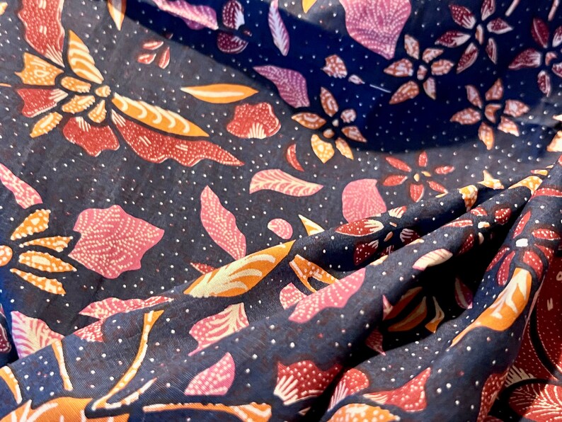 May include: A dark blue fabric with a repeating pattern of pink, orange, and white floral designs. The fabric is folded and draped, showing the intricate details of the pattern.