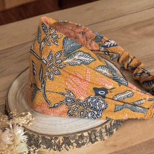 May include: Mustard yellow headband with a floral pattern in navy blue, peach, and white. The fabric is gathered at the back for a comfortable fit. The headband is displayed on a wooden surface.
