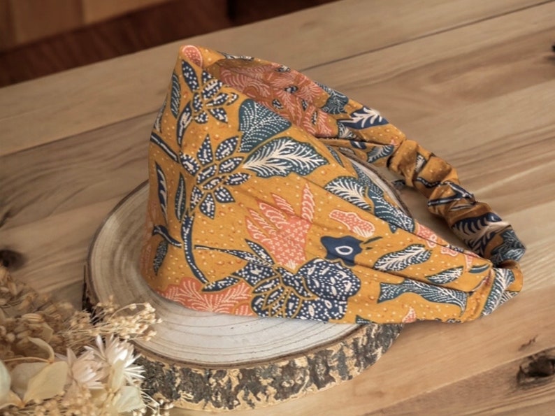 May include: Mustard yellow headband with a floral pattern in navy blue, peach, and white. The fabric is gathered at the back for a comfortable fit. The headband is displayed on a wooden surface.