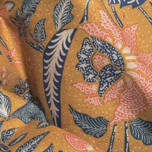 May include: Close-up of a fabric with a floral pattern. The design features large flowers and leaves in shades of pink, blue, and white against a mustard yellow background. The fabric has a textured appearance, with small dots throughout.