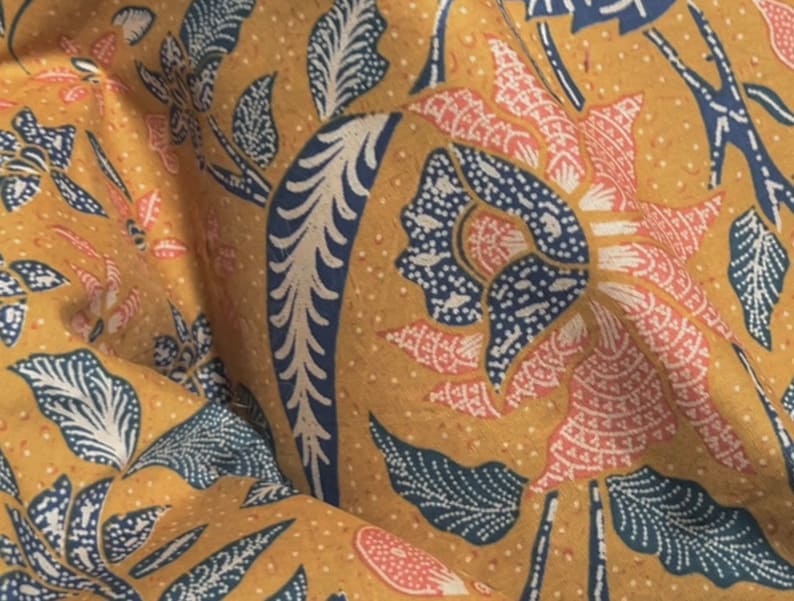 May include: Close-up of a fabric with a floral pattern. The design features large flowers and leaves in shades of pink, blue, and white against a mustard yellow background. The fabric has a textured appearance, with small dots throughout.