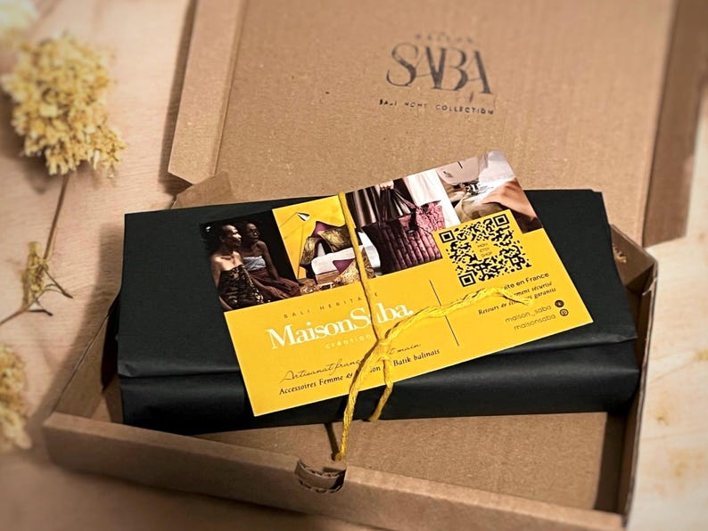 May include: A black gift box tied with a yellow ribbon and a yellow card with the text "Maison Saba" and a QR code. The card is on top of a brown cardboard box with the text "SBA Ball Home Collection".