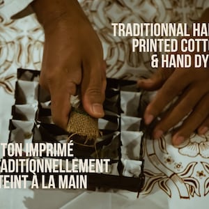 May include: A person's hands are shown using a traditional hand-printing technique on a white cotton fabric with a batik pattern. The text "TRADITIONAL HAND PRINTED COTTON & HAND DYED" is displayed above the hands. The text "COTON IMPRIM&Eacute; TRADITIONNELLEMENT & TEINT A LA MAIN" is displayed below the hands.