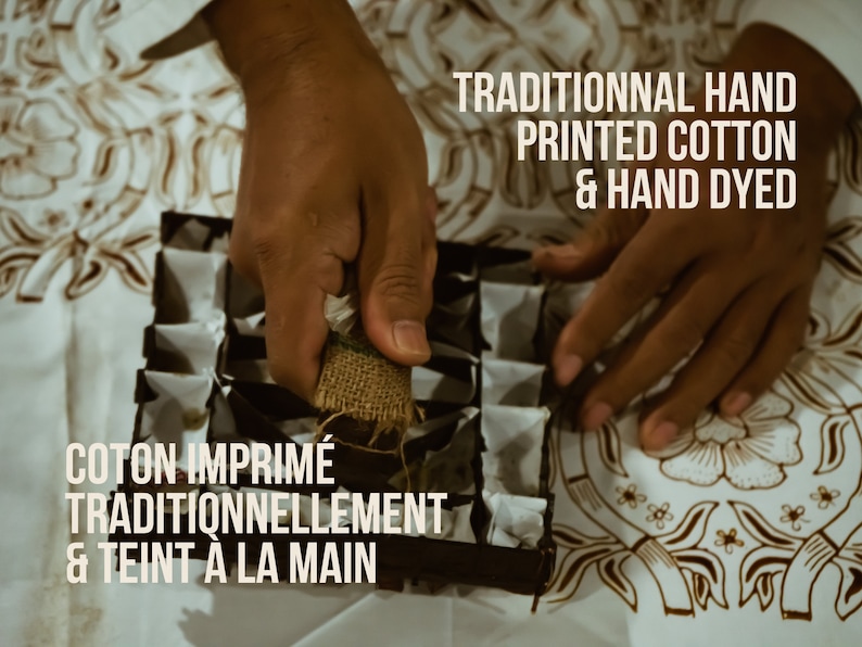 May include: A person's hands are shown using a traditional hand-printing technique on a white cotton fabric with a batik pattern. The text "TRADITIONAL HAND PRINTED COTTON & HAND DYED" is displayed above the hands. The text "COTON IMPRIM&Eacute; TRADITIONNELLEMENT & TEINT A LA MAIN" is displayed below the hands.